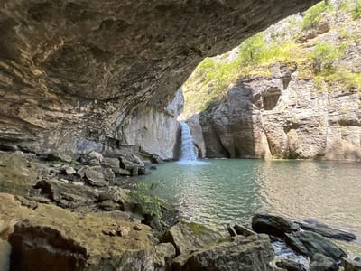 The image depicts a waterfall cascading into a pool of water within a cave, located in Emen, Bulgaria. The foreground is dominated by large rocks, some partially submerged in the water, leading to the pool. The cave's roof is visible above, textured with rock formations. A waterfall feeds the pool of water from the back of the cave. Outside the cave opening, rock walls with vegetation appear. The sunlight illuminates the rock walls outside the cave, in contrast to the relative shade within the cave's mouth.