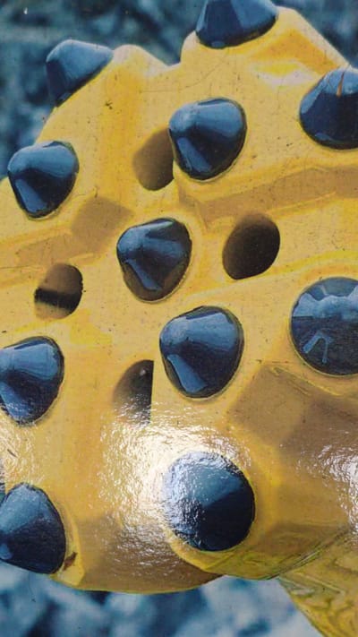 The image displays a close-up of a yellow, industrial drilling tool, likely a drill bit. The tool is characterized by its bright yellow, somewhat worn surface, showing fine cracks and speckles, indicating use. It features numerous dark, rounded protrusions embedded in its surface, which appear to be made of a darker material, possibly carbide or a similar hard substance, designed for drilling. These protrusions are arranged in a pattern across the tool's face, with some depressions or sockets also visible between them. The surface of the tool is somewhat glossy, reflecting light and highlighting its texture. The background is out of focus and appears to be a muted, grayish-blue, possibly suggesting a natural outdoor environment or a blurred industrial setting. There is no visible text in the image. The perspective is a low-angle shot, emphasizing the ruggedness and scale of the tool. The lighting suggests it might be daytime, possibly overcast given the diffused reflections, but this is not definitively ascertainable. The context of Goczałków, Poland, might relate to mining or construction activities where such a tool would be employed. There are no people or animals visible. The focus is entirely on the mechanical object.