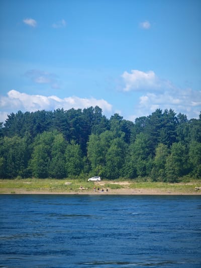 The image depicts a serene natural landscape with a wide, calm blue river in the foreground. The river stretches across the lower half of the frame, its surface textured by gentle ripples. In the middle ground, a sandy shore separates the river from a dense, lush green forest that dominates the upper portion of the image. The forest is a mix of deciduous and coniferous trees, with a varied canopy creating a rich tapestry of greens. A few scattered rocks are visible on the shore.
On the sandy shore, near the edge of the forest, a white van is parked. A small group of people, appearing to be enjoying the outdoors, are scattered along the shoreline. One person is standing near the water's edge, while others are seated or lying down, seemingly relaxing on the sand. The sky above is a bright, clear blue with a few scattered white cumulus clouds, suggesting a pleasant, sunny day. The overall impression is one of a peaceful, natural setting perfect for recreation, likely a riverside park or a natural area accessible by road, such as one might find near the settlement of Абалаково, Russia, given its proximity to the Yenisei River.