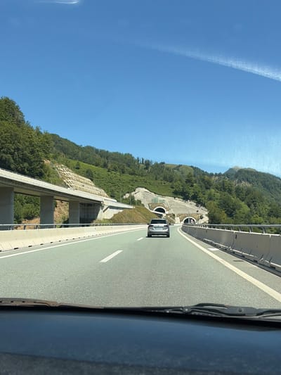 The image captures a highway scene in Lopate, Montenegro, on a clear, sunny day. The perspective is from inside a vehicle, looking forward through the windshield. The highway has multiple lanes, marked with white lines, and is bordered by concrete barriers on the right. In the distance, a silver car is traveling ahead. The road curves to the right, leading towards two tunnel entrances cut into a green, mountainous hillside. The hillside is covered with lush trees and features some constructed retaining walls with a grid pattern. A large overpass supported by concrete pillars is visible on the left side of the image. The sky is a bright blue with a faint, wispy contrail stretching across it.