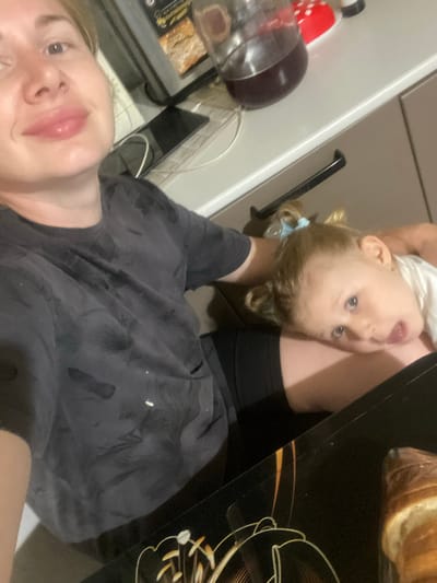 A woman, likely in her late twenties, with blonde hair and fair skin, is taking a selfie in what appears to be a kitchen. She is wearing a dark grey t-shirt with a camouflage pattern and black shorts. Her expression is neutral, with her lips slightly parted. A young child, with blonde hair tied in pigtails and blue eyes, is resting on the woman's lap, looking up at the camera with a surprised expression. The child is wearing a white t-shirt. The background shows a kitchen counter with a jar of dark liquid, a red bowl, and what appears to be a microwave. A dark-colored tray with a decorative pattern is in the foreground, and a loaf of bread is visible on the right side of the tray. The overall lighting suggests an indoor setting. The image does not contain any information that would allow for the determination of the location in the city of Маринопіль, Ukraine.