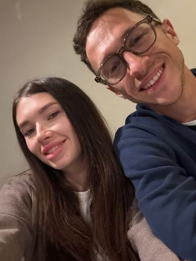 A close-up, tilted selfie features a smiling man and woman. The man, on the left, wears glasses and a blue hooded sweatshirt. The woman, positioned slightly lower on the right, smiles broadly with her tongue partially visible. Her dark, long hair cascades down her shoulder. Both individuals are looking towards the camera. The background appears to be a plain wall or ceiling, suggesting an indoor setting. The overall composition creates a casual and intimate snapshot.