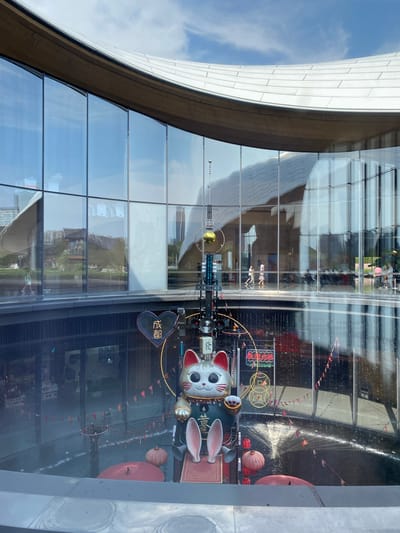 The image shows a circular open-air atrium in a modern building, likely a shopping mall or cultural center, located in Jinniu District, China. The building features a large, curved glass facade that reflects the sky and surrounding environment. Inside the atrium, a large decorative display is visible, centered around a statue of a Maneki-neko (beckoning cat). The cat statue is adorned in traditional Chinese attire and is positioned on a platform with red accents. Various other decorative elements are present, including red umbrellas, lanterns, and small triangular flags strung across the atrium. The reflections on the glass create a layered visual effect, showing people walking on an upper level of the building. The overall scene suggests a lively and festive atmosphere, possibly during a holiday or special event.