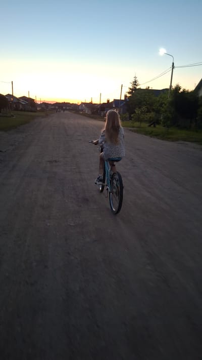 The image captures a young person riding a bicycle on a dirt road. The person has long, blonde hair and is wearing a light-colored, patterned shirt and dark shorts. They are positioned on the right side of the frame, cycling away from the viewer. The bicycle appears to be a medium-sized, light-colored model with black tires.
The scene is set in a rural or semi-rural area, likely in Belarus, given the provided location context. Houses with visible roofs and utility poles line the left side of the road, suggesting a village or small town. Dense greenery and trees are also present on the left, behind the houses.
The time of day is clearly indicated by the sky. The left side of the image shows a vast expanse of clear sky, transitioning from a pale blue at the top to a soft yellow and orange hue near the horizon, indicating either sunrise or sunset. This warm glow is reflected on the houses and trees. The overall lighting suggests either early morning or late evening. The weather appears to be clear, with no visible precipitation.
The dirt road is textured and appears somewhat uneven, with tire tracks visible. The overall mood is tranquil and serene, evoking a sense of peaceful outdoor activity during a beautiful part of the day. There is no visible text in the image.