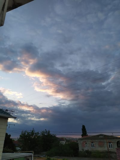 This is a photograph taken outdoors in Bilajari, Azerbaijan, likely during sunset given the dramatic cloud formations and the warm hues of orange and pink interspersed with dark grey. The image is oriented vertically, with the sky occupying the majority of the frame. Several power lines run diagonally across the sky, partially obscuring the view of the clouds.

In the upper right portion of the image, a building with a grey facade and arched windows is visible. The building appears to be two stories high with a pitched roof. Adjacent to the building, dense green foliage and trees are present, suggesting a suburban or semi-rural setting. In the background, a hazy vista of a city or town can be seen, further indicating a populated area.

The clouds are a prominent feature, with areas of bright light breaking through darker, more ominous clouds. This contrast creates a dynamic and somewhat moody atmosphere. No people are visible in the photograph. The overall impression is one of a picturesque natural phenomenon occurring over a developed landscape. There is no visible text in the image.