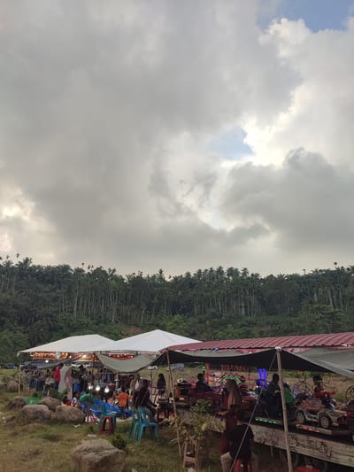 This image captures an outdoor market scene under a cloudy sky, possibly in Pantonrayeuk Satu, Indonesia. In the foreground, several stalls are set up under awnings, with people browsing and interacting. One stall appears to be selling food or drinks, with lights illuminating the offerings. Another stall features a small amusement ride with several children seated in toy vehicles. The ground is covered in grass and scattered with large rocks. Behind the market, a dense line of trees, primarily tall, slender palms, forms a backdrop against the hilly terrain. The sky is dominated by heavy, grey clouds, with a small patch of blue visible in the upper right quadrant. The overall atmosphere suggests a local gathering or festival taking place in a rural or semi-rural setting.