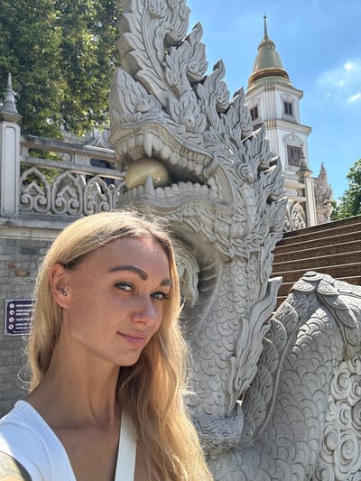 A woman with blond hair stands in front of a large stone dragon statue, with a temple structure visible in the background. The location is Dĩ An, Vietnam, as indicated by the Vietnamese language sign on the lower right. The woman is wearing a white top and has a piercing in her ear. The temple is intricately designed with a tiered roof structure. Trees are visible, suggesting a partially outdoor setting. The woman is facing the camera while holding the camera herself.
