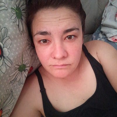 The image is a close-up, overhead shot of a woman lying down. She is wearing a black tank top. Her face is directed towards the camera, and she has a neutral expression. She has brown eyes and dark hair. Her skin appears to have some redness. To her left, there is a pillow with a pattern of white daisies on a grey background. To her right, there are gray blankets or other bedding. The lighting suggests it might be daytime, possibly indoors due to the soft illumination. The context provided is Podlubovo, Russia. There is no visible text in the image.