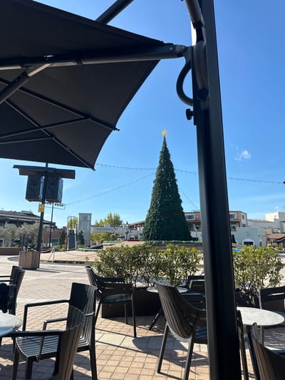 The image depicts an outdoor public or commercial plaza in Marratxí, Spain, under clear daylight conditions. In the immediate foreground, the frame is partially occupied by a large, open, black patio umbrella on the left and its structural pole on the right. Below the umbrella, several unoccupied dark-toned outdoor chairs, some with slatted backs, are arranged around small, round, light-colored tables on a reddish-brown brick-paved ground. In the midground, a tall, conical artificial Christmas tree, dark green and topped with a golden star, stands centrally, encircled by a low green hedge. Horizontal string lights are strung across the plaza. To the left of the Christmas tree, a light pole with two black speaker units is visible. In the background, buildings featuring light-colored facades and tiled roofs are present, along with an escalator structure and a sign labeled "RESTAURANTES." No individuals are depicted in the scene.