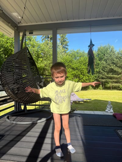 A young boy stands on a dark wooden porch with shadows cast by sunlight. He wears a pale yellow t-shirt with text that reads "MAKE THINGS HAPPEN" and dark blue socks with white accents. The boy has his arms outstretched, palms facing forward, and is looking down and to his right. To his left is a large, dark woven egg-shaped hanging chair. Behind the boy, a green lawn is visible, dotted with trees and a bright blue sky. A decorative witch figure hangs from the porch ceiling, and a small stone statue of a meditating figure sits on the edge of the porch. The scene is likely a residential backyard in the city of Алешкино Дп, Russia.