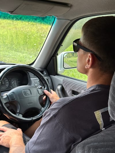 A person is driving a car, seen from the passenger's perspective. The driver, wearing sunglasses, has their hands on the steering wheel. Outside the car, a grassy landscape is visible. The dashboard and interior of the car are also in view. The location is the city of Nalobino, Russia.