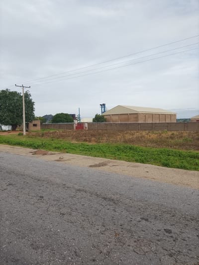 The image shows an overcast sky above a rural-looking scene in Barikin Sibre, Nigeria. In the foreground, a grey asphalt road with some cracks and patches runs diagonally across the bottom of the frame. To the right of the road, a strip of dry, yellowed grass and some green vegetation lines the edge.
Further back, a fence made of concrete blocks with barbed wire on top encloses a property. Behind the fence, there is a long, light-colored industrial building with a sloped roof, possibly a warehouse or factory. To the left of this building, part of a blue industrial structure is visible. Adjacent to the fence, there are some smaller structures, including a building with a small window and a red gate.
To the far left of the image, a large, leafy green tree stands next to a utility pole with wires extending across the sky. The overall environment appears to be a transition zone between undeveloped land and an industrial area. No people are visible in the image.