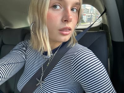 An individual with blonde hair and light-colored eyes is depicted inside a vehicle, secured by a seatbelt, looking directly at the camera. The subject wears a horizontally striped navy blue and white long-sleeved shirt, large silver hoop earrings, a nose ring on the left nostril, and multiple necklaces, including one with a silver starfish pendant. Her mouth is slightly open, revealing the tip of her tongue. The vehicle's dark-colored interior and seat are visible. Through the rear window, an urban street scene is observed. A multi-story building with distinct windows and architectural features is prominent in the background. Signage on this building clearly identifies it as "HARPO'S BEER & WINE" or "HARPO'S RESTAURANT." This building is historically located at 630 Johnson Street, Victoria, British Columbia, Canada.