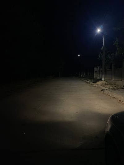 The image depicts a nighttime scene on a paved road in Slobozia-Dușca, Moldova. A street light illuminates a portion of the road, casting long shadows. The road extends into the darkness, bordered by a fence and some foliage on the right side. In the foreground, the edge of a dark-colored vehicle is visible on the right, with its headlights casting a beam onto the road. The environment appears to be a quiet, possibly suburban or rural area, with limited visible human activity.