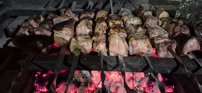 In Molovata Nouă, Moldova, an outdoor barbecue features skewers of meat cooking over glowing red embers. The meat, cut into large cubes, is seasoned and shows signs of browning and charring on the exterior. Several metal skewers are loaded with the meat, arranged in rows on a dark metal grill. The background is dark, with the bright, intense light of the coals illuminating the underside of the skewers and the meat. The overall scene captures a moment of food preparation in a rustic setting.