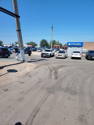 The image depicts an outdoor scene on a sunny day in Тораңғылық, Kazakhstan. In the foreground, a paved surface shows tire tracks and dark stains, possibly oil or tar. A shadow of a person is visible on the lower left. In the mid-ground, a concrete utility pole stands to the left, with wires running across the frame. Two individuals are walking away from the camera on the right side of the pole. One, a male wearing a black t-shirt, grey sweatpants, and a baseball cap, carries a blue bag over his shoulder. The other, a female wearing a black top and skirt and carrying a backpack, walks with bare feet.
Behind the individuals, a parking lot is visible with numerous cars. To the right, a building with a blue facade and white lettering that reads "POLISIA" is prominent. Adjacent to the "POLISIA" building, other structures and shops are visible, including one with red signage featuring Cyrillic lettering that appears to be a "Doner" shop. The background consists of trees and a clear blue sky. The overall environment suggests a commercial or service area within a town or city.