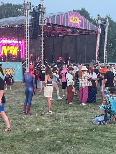 A wide shot captures a lively outdoor event, possibly a music festival, in Surakovo, Russia. The scene is dominated by a large stage structure with prominent audio equipment and bright pink and purple lighting illuminating the upper canopy. A large LED screen displays vibrant yellow text in Russian, possibly the event's name. In the foreground, a crowd of people are gathered on a grassy field. Among them, a person dressed in a full Spider-Man costume stands out, facing away from the camera. Other attendees are seen in casual attire, some conversing, others looking towards the stage. A woman in a green top and white shorts is visible to the right of the Spider-Man figure. Further into the crowd, individuals wear various hats, and some carry bags. The environment appears to be a park or open field, with trees visible in the background. The overall atmosphere suggests a communal gathering, likely enjoying a performance or event.