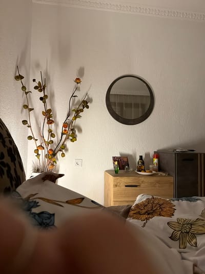 The image depicts a bedroom scene, likely in Zmeitsa, Bulgaria. In the foreground, a person's hand and part of their arm are blurred, suggesting they are taking the photo from a bed with floral-patterned bedding. The bed covers feature large yellow and blue floral designs.

In the mid-ground, a wooden nightstand is visible. On top of the nightstand are several items: a framed photograph of people, a small bottle of green liquid, a small clear bottle, a white dish with fruit, and two aerosol cans, one red and one yellow. To the right of the nightstand is a dark grey cabinet.

Further back, a white wall serves as the backdrop. A circular mirror with a dark frame hangs on the wall above the nightstand. To the left of the nightstand, a decorative branch with artificial flowers and leaves is placed in a white vase. A white electrical outlet is visible on the wall near the vase. The room is lit by artificial light, casting shadows on the wall.