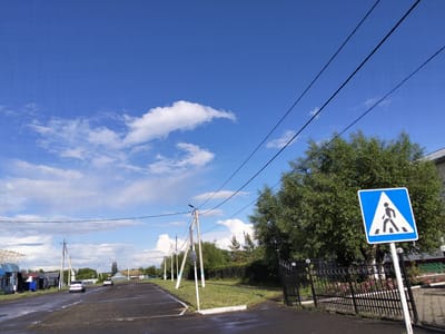 This image depicts a street scene in Bulaev, Kazakhstan, under a bright blue sky with scattered white clouds. The foreground features a wet asphalt road with several puddles. To the right, a pedestrian crossing sign, a blue square with a white triangle containing a black silhouette of a person walking, is mounted on a white pole. Behind the sign is a black metal fence with decorative elements and a dense green tree. The midground shows a row of utility poles with overhead wires stretching across the scene. Several houses and other buildings are visible on the left side of the road, some with corrugated metal roofs. Two cars, a silver sedan and a white SUV, are driving on the road. The background consists of more trees and houses, with a slightly hazy horizon. The overall impression is of a semi-urban or rural environment on a sunny day.