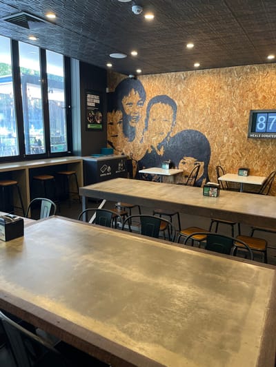 This image captures the interior of a modern, fast-casual dining establishment, likely a restaurant or cafe, seen from an aisle between tables.
The ceiling, located on the left side of the frame, is dark grey with a textured, patterned finish, illuminated by several circular recessed lights.
A dominant feature is the main wall, constructed from light-colored oriented strand board (OSB) or particleboard. A large, black stenciled mural depicting the faces of two individuals in a graphic style adorns this wall. Above the mural, a digital display shows the number "87" and the text "MEALS DONATED," indicating a charitable initiative.
In the foreground and mid-ground, two long, thick communal tables with smooth, light grey, concrete-like surfaces extend through the space. Arranged along these tables are various chairs and stools; some chairs feature dark green backs and wooden seats, while others are simple bar stools. Small, white table tents are visible on some of the smaller tables positioned closer to the wall.
Towards the lower left, a large window spans the height of the wall, allowing ample natural light into the space and offering a view of outdoor greenery, including trees. Below the mural, on a dark wall section, a framed poster advertises the "CHILDREN'S CHEESEBURGER PROJECT," with additional text that is not clearly legible. Further to the right, waste sorting bins are marked "GENERAL WASTE" and "BOTTLES + CANS."
No individuals are present in the image, suggesting a quiet time, possibly outside of peak operating hours. The natural light streaming through the window indicates it is daytime. The overall aesthetic combines industrial and rustic elements, with a clear emphasis on communal dining and a charitable aspect evident from the "MEALS DONATED" display and the "CHILDREN'S CHEESEBURGER PROJECT" poster. The location context provided is Kelvin Grove, Australia.