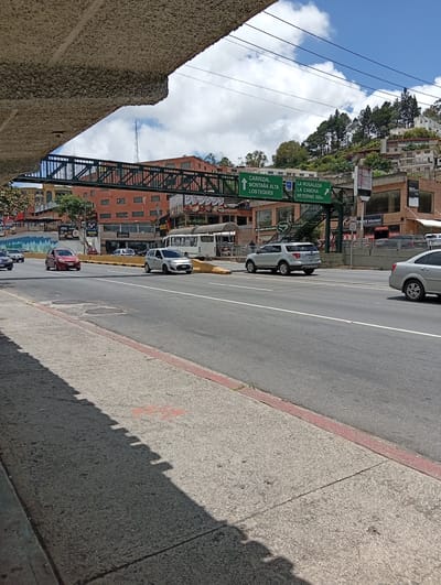 The image shows a street scene in Carrizal, Venezuela. A multi-lane road runs horizontally across the frame, with several cars driving on it. On the left side of the road, a pedestrian bridge spans across the lanes. Buildings are visible behind the bridge and along the side of the road, some with signs. A hillside covered in trees and houses rises in the background. The sky is blue with scattered white clouds. A shadow of an overhead structure falls across the sidewalk in the foreground.