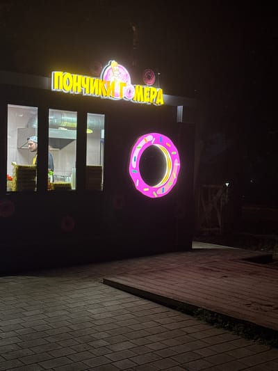 A neon sign with yellow lettering reads "ПОНЧИКИ" which translates to "donuts" in Russian. The sign also features a large pink neon donut with colorful sprinkles, and a smaller donut shape next to the lettering. The sign is illuminated at night, casting a glow on the dark facade of a building. Behind the sign, three rectangular windows reveal the interior of a donut shop. In the windows, kitchen equipment and shelves stocked with donut boxes are visible. In the bottom window, two individuals are seen working. The scene is set outdoors, with a brick wall visible on the right side of the image. The overall impression is a nighttime shot of a brightly lit donut shop in Vladikavkaz, Russia.