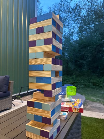 This image depicts a large Jenga tower constructed from wooden blocks of varying colors, including natural wood, light blue, and purple. The tower is precariously stacked on a dark-colored outdoor table. In the background, a green corrugated metal wall is visible to the left, and trees with a twilight sky can be seen to the right. A portion of a blue cushioned chair is visible in the lower left corner. On the table, to the right of the Jenga tower, there are several items, including a blue lattice container, a bright green lattice container, and a board game box that appears to be "Guess Who?". The scene is set outdoors, likely in a backyard or patio area, given the furniture and natural surroundings. The lighting suggests it is dusk. This scene is located in the Town of Dell Prairie, United States of America.