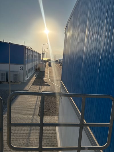 The image depicts an exterior view of a modular building complex bathed in the warm, low light of a setting or rising sun. The sun's glare creates a strong lens flare diagonally across the upper portion of the image. In the foreground, a metallic railing suggests a viewing platform or walkway.
To the left, a long, two-story modular building with white lower walls and blue upper walls is visible. Its facade features numerous windows. Several large, rectangular air conditioning units are positioned at the base of the building. The number "90 VRF 02" is clearly marked on the building's exterior. A paved walkway of interlocking concrete blocks runs alongside this building.
Further down the paved area, a road is visible, with streetlights lining its edge. Several trucks and other vehicles are present on the road, suggesting an industrial or logistical area. In the distance, a mountainous or hilly landscape is faintly visible against the bright sky.
To the right, a tall, blue, corrugated metal structure dominates the view, its ribbed surface reflecting the sunlight. This structure appears to be another modular building or a large storage unit. The overall scene suggests a modern, functional facility, possibly an administrative or accommodation complex within an industrial zone or port. The setting is likely outdoors, with evidence of infrastructure such as roads, lighting, and paved pathways. The location is identified as the city of Gülnar, Turkey.