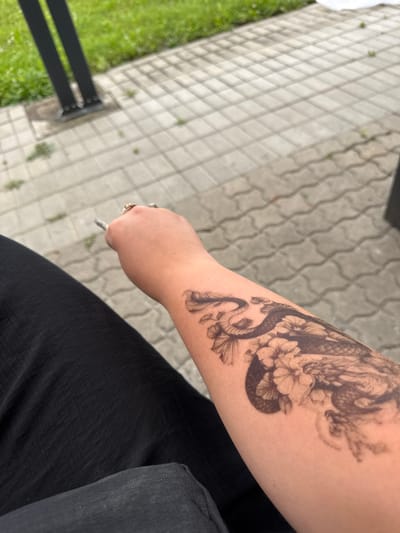 An arm with a tattoo of a dragon and flowers is held up, with a cigarette in the person's hand, in what appears to be an outdoor setting. The arm is resting on dark fabric, likely clothing. The background consists of light-colored paving stones and a patch of green grass. Two dark vertical posts are visible in the upper left quadrant of the image. The lighting suggests it is daytime. The location is identified as Bad Säckingen, Germany.