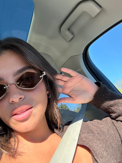 The image portrays a female individual seated inside a vehicle, reportedly in Lawrence Township, United States, under a clear, bright blue sky. The subject, positioned in the foreground, exhibits long dark brown hair and light brown skin, with glossy lips. She wears angular, silver-framed sunglasses with dark lenses. Her attire includes a brown, long-sleeved top or sweater, and a light-colored seatbelt is fastened diagonally across her chest and shoulder. Her right hand is raised, with fingers near her right temple, appearing to touch her hair. The vehicle's interior features a light-colored headliner and an overhead grab handle. The external environment, visible through the windows and reflected in the sunglasses, consists of a clear blue sky and green deciduous foliage.
