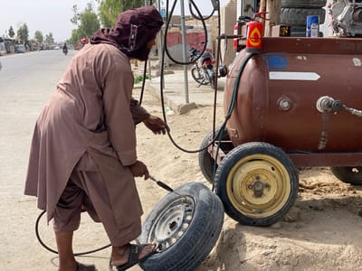 A man wearing a brown robe and a brown head covering is inflating a tire with an air compressor. He is standing in a dirt lot in Panjwai, Afghanistan, a rural area located at the coordinates 31.542, 65.463. He is using a hose to fill the tire. Behind him is a large brown water tank with a hose, a fire extinguisher, and other hoses. The tank is mounted on a metal frame with wheels. The scene is shot from the perspective of someone standing behind and to the right of the man. The air compressor is visible in the foreground. The man's right hand is holding the hose, and his left hand is on the tire. He appears to be focused on his task. The ground is a mix of dirt and gravel. The background is out of focus. The day is clear and sunny. There is a motorbike parked to the left of the tank. The man is the only person visible. The scene suggests that the man is working on a vehicle or a machine. The scene is peaceful and quiet.