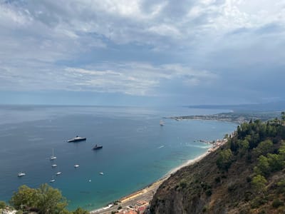 This is a high-angle, medium-shot photograph of the coastline near Taormina, Italy, on a cloudy day. The scene is dominated by the blue-green sea on the left, meeting a sandy beach and a cliffside town on the right. In the foreground, a steep, dry, vegetation-covered cliff drops sharply towards the sea. Small buildings with red-tiled roofs are clustered at the base of the cliff. Above the beach, a railway line runs parallel to the coast, with some low-rise buildings situated next to it. Further inland, the town of Taormina stretches along the coastline, characterized by its white buildings and abundant greenery. Several boats and yachts are visible on the water, including two large, dark-colored superyachts and numerous smaller sailing boats and speedboats. The sea is calm, with a clear demarcation between the deeper blue waters and the shallower, turquoise areas near the shore. The sky is filled with scattered, wispy clouds, with patches of brighter sky peeking through. The overall impression is of a scenic coastal landscape with evidence of human settlement and maritime activity.