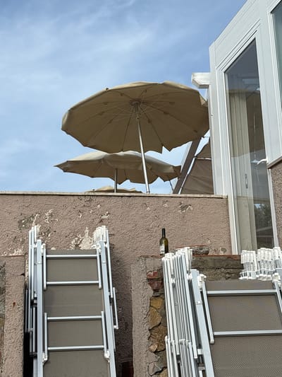 This media file captures an outdoor scene, likely in Portoferraio, Italy, featuring a building facade and stored beach equipment under a clear blue sky.

The main subject is a light brown, textured building wall, which exhibits signs of wear with areas of peeling paint and visible stonework at its base. A white-framed window or door, with glass panes reflecting some light, is visible in the upper section of the wall.

Below the window, a green wine bottle, possibly Chianti in shape, is horizontally positioned within a crevice or opening in the wall. Its white label is partially visible, but the text is illegible.

To the right of the wall and extending towards the bottom, numerous white-framed beach chairs or loungers are neatly stacked. These chairs feature grey, textured mesh fabric seats and backs. Their organized arrangement suggests they are in storage, perhaps before opening for the day, after closing, or during an off-season period.

On the left side of the image, contrasting with the building, several beige beach umbrellas are visible against the bright, clear blue sky. They appear to be partially open or folded, with their metal frames and fabric canopies clearly discernable.

The scene is devoid of human presence, conveying a sense of quiet or dormancy. The bright sky indicates clear daylight and favorable weather conditions. The presence of stacked beach furniture and umbrellas strongly suggests a setting in a coastal or resort area.