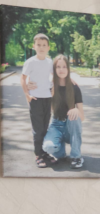 This is a photograph of two young people, likely siblings, posing outdoors. The person in the foreground, appearing to be a boy, is wearing a white t-shirt and dark pants. He is looking towards the camera with a neutral expression. Behind him, a girl is embracing him from behind, her arms wrapped around his torso. She has long brown hair and is wearing a dark long-sleeved shirt and blue jeans. She is also looking towards the camera with a slight smile.

The setting appears to be a park or a residential area with lush green trees and foliage in the background. The ground is paved, suggesting a pathway or sidewalk. Sunlight filters through the leaves, casting dappled shadows on the ground, indicating it is daytime, likely sunny. The overall mood of the photograph is casual and affectionate, capturing a moment between two young people. No text is visible in the image. The location context is provided as Berestyn, Ukraine.