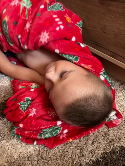 A young child is sleeping, wrapped in a red blanket with a Christmas pattern. The child's head is resting on a beige carpet, and a wooden surface is visible in the background. The scene appears to be indoors.