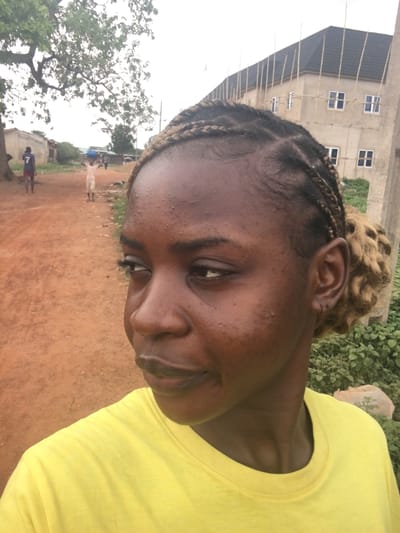 The image captures a slightly tilted, close-up outdoor portrait of a young person, likely a woman, with dark skin and a gentle, subtle smile. She is wearing a bright yellow t-shirt. Her hair is styled in cornrows with a bun of lighter, curly hair at the back of her head, just above her right ear.
The setting appears to be a rural or semi-urban area in Enyibichiri, Nigeria. The foreground and mid-ground consist of reddish-brown dirt or earth. In the background, there are various elements: a large tree with sparse foliage on the left, and a multi-story building on the upper left, which appears to be under construction or renovation, featuring visible scaffolding and several rectangular windows. Greenery and bushes are scattered around the environment.
Further in the distance, on a dirt path, several indistinct figures are visible. One figure stands out wearing a blue shirt with the white number "7" clearly printed on the back. The sky is overcast, providing soft, diffused natural light to the scene. The image conveys a calm, everyday moment in an outdoor local setting.