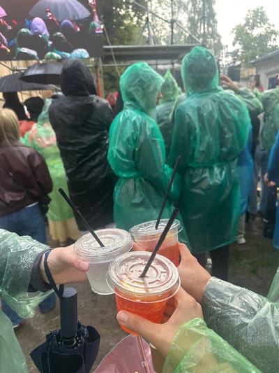 In Ivano-Frankivsk, Ukraine, three individuals, shielded by green rain ponchos, are toasting with drinks in clear plastic cups. Two of the cups contain a reddish-orange liquid with bubbles, and one contains a clear liquid. Black straws are visible in each cup. In the foreground, a partially closed black umbrella rests against the arm of one of the individuals. The background shows a crowd of people, many also wearing rain ponchos in green and black, with some holding umbrellas overhead. The environment appears to be an outdoor event, possibly a concert or festival, with a large screen visible in the upper left corner displaying illuminated shapes and people holding up phones. The weather is overcast and rainy, indicated by the wetness on the ponchos and the presence of umbrellas.
