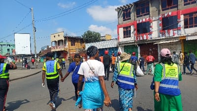 The image presents a daytime street scene in Gisenyi, Rwanda. A group of approximately ten individuals is walking forward on a paved asphalt road, moving away from the camera's viewpoint. Six individuals in the foreground and mid-ground are wearing high-visibility vests composed of yellow-green and blue panels.

From left to right in the foreground:
1.  An individual wears a red cap, dark attire, a high-visibility vest, and white gloves, with the right arm raised.
2.  A male in a dark cap, a yellow-green and blue high-visibility vest, dark track pants, and sneakers, is holding hands with another person in a blue shirt (partially obscured).
3.  A female wears a dark patterned head wrap, a white t-shirt with a graphic on the back, a light blue fabric tied around her waist, and dark patterned pants.
4.  A female wears a wide-brimmed khaki hat, a blue and white patterned long dress, and a yellow-green and blue high-visibility vest.
5.  A female wears a pink baseball cap, a green top, and a yellow-green and blue high-visibility vest with partially legible white text, including "UMUGANDA".

Numerous other pedestrians are visible further down the street, some carrying white bags.

The urban environment includes various multi-story buildings. On the right, a three-story concrete building features upper-floor windows with red frames and peeling white surrounds, and ground-level shopfronts with closed green and brown roll-down shutters. On the left, a large, blank white billboard stands adjacent to a wooden utility pole with multiple wires. Further left, buildings with light green/blue and orange/brown facades are present. The sky is blue with scattered white clouds.
