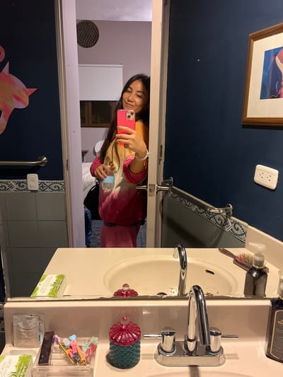 A woman is taking a selfie in a bathroom mirror. The mirror is above a sink with a chrome faucet and a variety of toiletries. The bathroom has blue walls with decorative tiles and a painting or print on the wall. There are also light switches and an outlet visible. The woman is wearing a pink and white sweater and holding a water bottle in one hand and her pink phone in the other. The bathroom has light grey tiles. The exact location in the city of Santiago de Surco, Peru cannot be determined from the image alone.