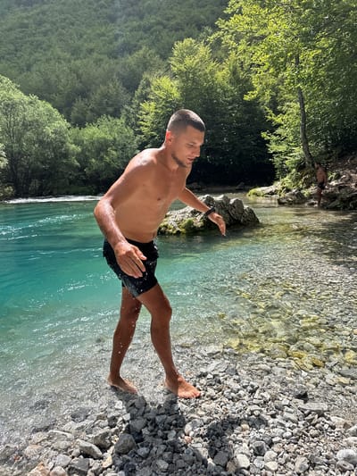 A man is standing in a shallow stream in Vusanje, Montenegro. He is shirtless and wearing black swim trunks. His body is wet, suggesting he has just entered the water or is about to. The stream has clear, turquoise water and a rocky bottom. Behind the man, the stream flows into a wider body of water, and the banks are covered in lush green trees. Sunlight filters through the leaves of the trees, dappling the water and the ground. In the background, another person is visible walking along the bank of the stream. The overall scene depicts a natural, serene environment.