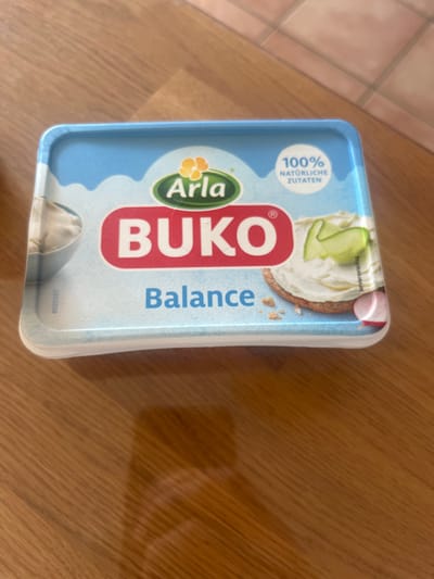 The image shows a container of Arla Buko Balance spread, sitting on a wooden table surface.  The container is blue, white, and red and features the Arla and Buko logos as well as the words "Balance" and "100% naturliche Zutaten". The spread contains white cream cheese, with a green cucumber slice visible on top. The image was taken in the city of Köln, Germany, at the coordinates 50.934 latitude and 6.974 longitude.