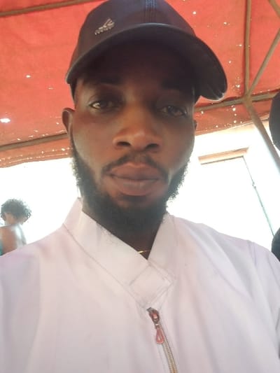 A close-up photograph, taken in the city of Ochadam, Nigeria, features an adult male positioned centrally. He has dark skin, dark eyes, and a trimmed beard, looking directly at the camera with a neutral expression. He wears a dark baseball cap with a light-colored emblem visible on the front and a white collared jacket secured by a zipper with a distinct red pull tab. A thin gold chain is visible around his neck beneath the jacket collar. In the blurred far-left background, the back of a head with dark, curly hair is partially discernible, possibly belonging to a child. The upper background shows a red or reddish-orange canopy structure with visible metal support poles, indicating an outdoor setting. The remainder of the background is brightly illuminated and out of focus.