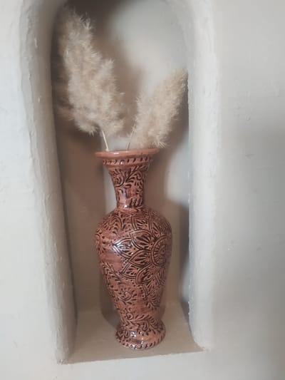 A tall, decorative ceramic vase sits on a shelf within a recessed niche in a wall. The vase is a reddish-brown color with intricate black patterns of swirling vines, leaves, and circular motifs adorning its surface. Two sprigs of dried, feathery beige grass are placed inside the vase. The niche is softly lit, with the rough texture of the wall visible. The overall aesthetic suggests a traditional or rustic interior design, potentially found in a historic building in Khiva, Uzbekistan, known for its preserved ancient architecture.