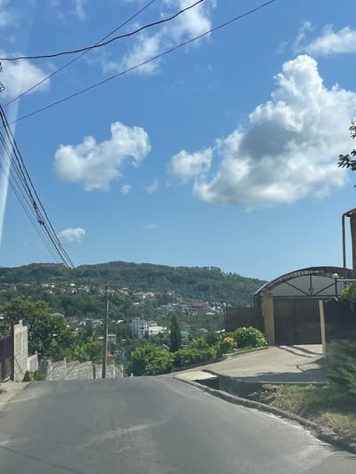 The image depicts a view of a residential area on a hillside under a bright blue sky with scattered white clouds. A paved road curves towards the viewer from the foreground. To the left of the road, a concrete wall separates it from a property with a metal gate. Further back, a densely wooded hillside is dotted with numerous houses and buildings of various sizes and architectural styles, with some appearing to be multi-story. Power lines are visible stretching across the upper portion of the frame. On the right side of the image, a large, arched gate stands in front of a property. The overall impression is of a sun-drenched, semi-urban environment nestled within a verdant, hilly landscape. This scene is located in the village of Verkhniy Yurt, Russia.