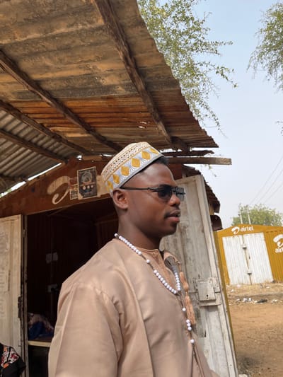 The image depicts an outdoor scene in Kanti, Nigeria, under bright daylight. A young adult male is positioned in the foreground, seen in profile, looking towards the right. He possesses a medium-dark complexion, wears dark sunglasses, a light brown traditional garment, and a white and gold embroidered kufi cap. A long white beaded necklace with two tassels is draped around his neck. Behind the individual, to the left, is a weathered, small wooden structure, likely a stall or shop, featuring a rusty corrugated metal roof supported by wooden beams. Faded posters are visible on the structure's wall, and an open doorway reveals a dimly lit interior. To the right in the background, a yellow structure, possibly a gate or kiosk, displays the red "Airtel" logo. The ground is unpaved earth, and sparse tree foliage is visible against a clear blue sky.