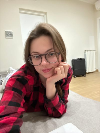 This indoor scene, reportedly located in Scafati, Italy, depicts a female subject lying prone on a grey upholstered surface. The subject has medium-length brown hair and wears dark-rimmed glasses along with a red and black plaid button-up shirt. She is propped on her left elbow, with her right hand resting under her chin, and gazes directly forward towards the camera with a slight smile. The background features light-colored walls, a white interior door, and light wood flooring. To the right, a white wall-mounted radiator is visible, positioned next to a dark-colored, four-wheeled hard-shell suitcase. A small, rectangular, reflective silver art piece is mounted on the wall to the left of the door. A portion of a white electronic device is partially visible in the lower right foreground.