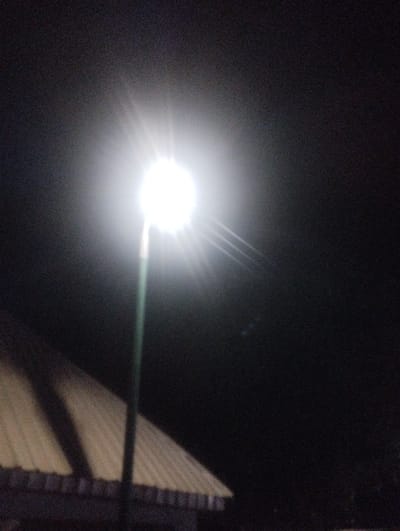 In the City of Healthcare, Nigeria, an intensely bright artificial light source, likely a streetlamp, is centrally positioned, emitting a prominent starburst effect against a dark night sky. A slender, dark pole supports the light, extending vertically. A section of a light-colored, corrugated metal roof, featuring parallel ridges and sloping downwards to the right, is visible in the lower-left quadrant. The overall environment is dark, exhibiting digital noise, and no individuals or specific activities are discernible.