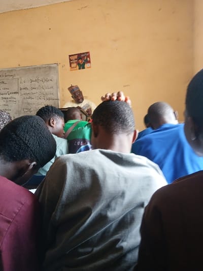 The image displays an indoor scene within a building in the city of Ugi Majowel, Nigeria. The room features light brown/beige walls, with a whiteboard on the left displaying equations and text written in white. A small, colorful poster is attached to the wall to the right of the board. In the foreground, multiple individuals are visible from behind, suggesting they are seated or standing as an audience. These include a person wearing a dark maroon shirt on the far left, an individual in a light olive-green long-sleeved shirt in the center, and another in a blue shirt to the right. In the background, an adult male with a beard is visible, wearing a light-colored patterned cap and a light-colored, possibly cream, traditional garment. He is holding a stack of white rectangular objects, consistent with papers or currency, in his hands, looking down at them. The individuals in the foreground are oriented towards this male figure.