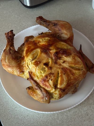 A whole roasted chicken sits on a white plate, which rests on a speckled grey countertop. The chicken is golden brown and appears to be seasoned with herbs. The background shows a blurred kitchen appliance. The image is taken from a slightly elevated angle.
