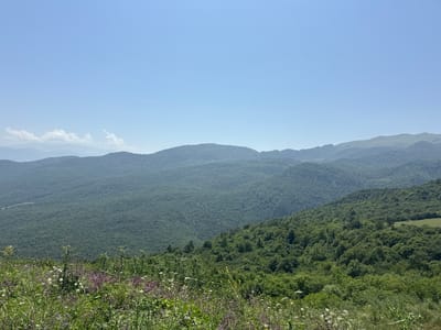 The image shows a panoramic view of a mountainous landscape in Yenokavan, Armenia. In the foreground, there is a field of wildflowers and tall green grass. The middle ground is dominated by rolling hills covered in dense green forests. In the background, more distant mountains can be seen, fading into a hazy blue sky. The sky is clear with a few scattered white clouds. The overall impression is one of natural beauty and serenity.